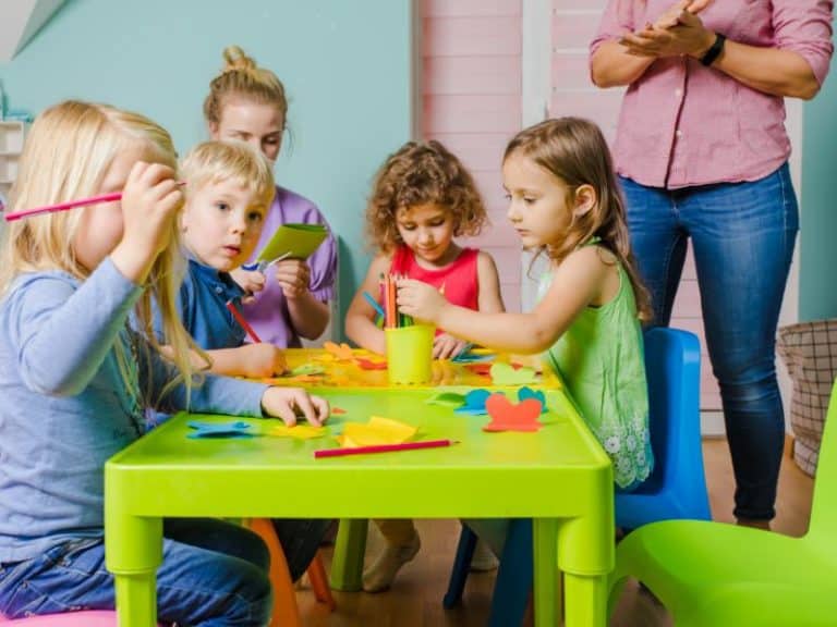 crianças em volta de uma mesa pedagógica e uma cuidadora ao fundo monitorando as crianças