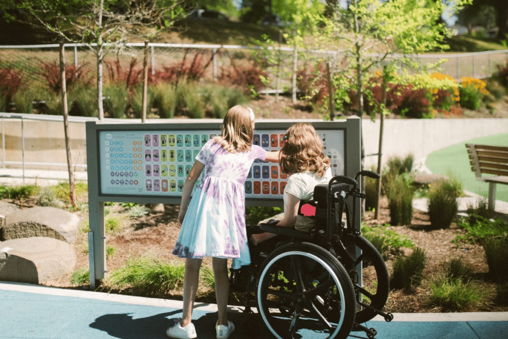 Duas crianças, uma delas em cadeira de rodas, interagindo com um painel educativo em um parque acessível e inclusivo, rodeadas por vegetação e áreas de lazer.