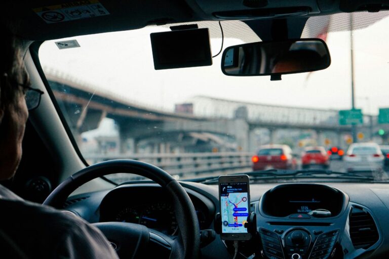 Motorista conduzindo um veículo em uma via movimentada, com um aplicativo de navegação aberto no celular preso ao painel. A imagem reflete a prática da direção defensiva, com o motorista atento ao trânsito à frente, utilizando tecnologia para planejar sua rota de forma segura.