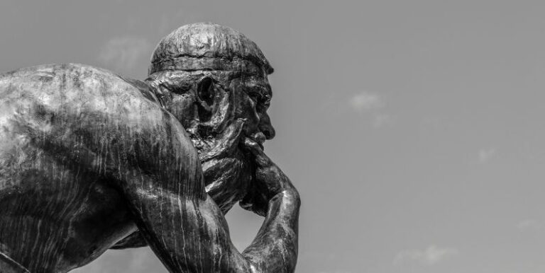 Escultura metálica de um homem idoso e musculoso em posição pensativa, com a mão no queixo e olhar introspectivo. A imagem em preto e branco destaca os detalhes da textura do metal e os traços fortes do rosto da estátua, contrastando com o céu nublado ao fundo. A cena evoca reflexão e contemplação filosófica.