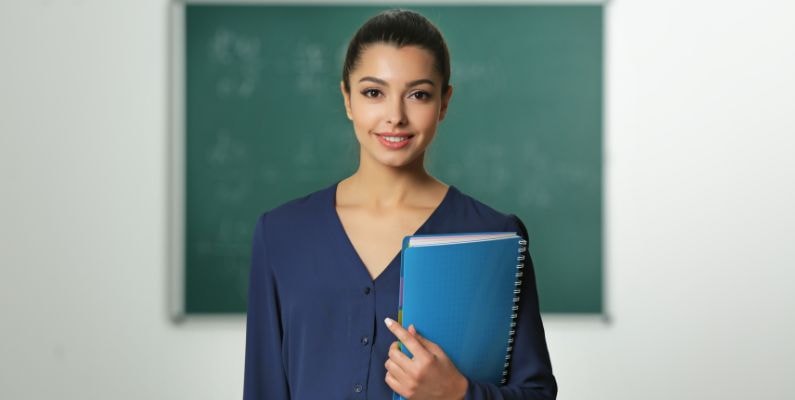 Uma professora sorridente em frente a uma lousa verde, vestindo uma blusa azul marinho e segurando um caderno azul. O fundo mostra uma sala de aula com algumas anotações na lousa. Ela tem o cabelo preso e transmite uma aparência profissional e acolhedora.