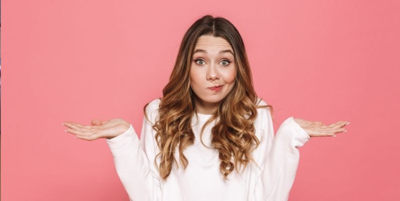 Mulher jovem de cabelos longos e ondulados, usando uma blusa branca, com expressão facial de dúvida e braços levantados com as palmas das mãos voltadas para cima, como se estivesse indecisa ou não soubesse a resposta. Fundo rosa sólido.